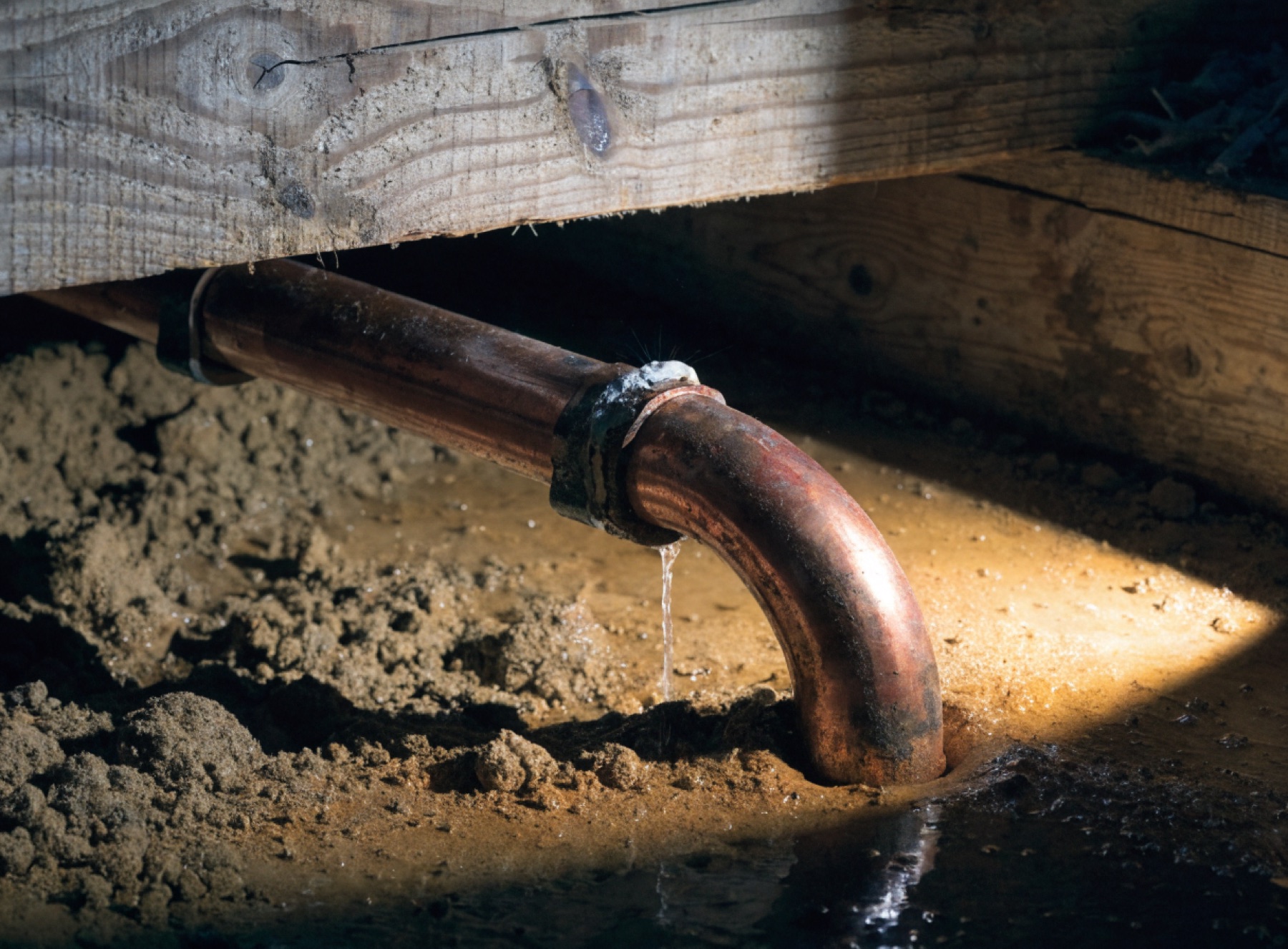 A copper water pipe in a subfloor crawl space with water spraying from a failed joint